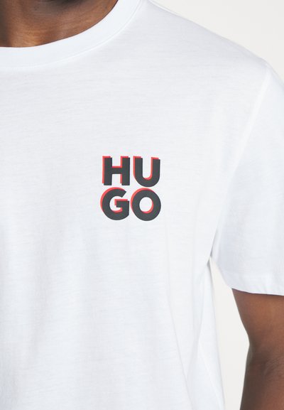 T-shirt en coton blanc avec un graphique sur la poitrine gauche. Le texte "HU" est rouge et "GO" est noir, avec une texture légèrement en relief. Manches courtes.