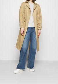 Beige Trenchcoat mit großen Knöpfen, kombiniert mit einem hellgrauen Pullover, weiten blauen Jeans und weißen Sneakers.