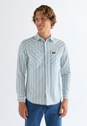 Mężczyzna ubrany w jasnoniebieską, długorękawową koszulę zapinaną na guziki z białymi pionowymi paskami i dwoma przednimi kieszeniami, w połączeniu z niebieskimi dżinsami.