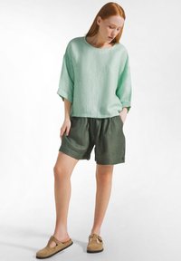 Vrouw met een lichtgroene loszittende top en donkergroene korte broek, staand met één hand in de zak en naar beneden kijkend, beige instapschoenen.