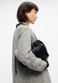 Bolso negro con pliegues sostenido contra un suéter de punto gris claro, mostrando un asa redonda y cierre de cuerda. Diseño simple y texturizado.