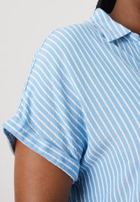 Ljusblå kortärmad skjorta med vita vertikala ränder, med en spetsig krage och uppprullade ärmkanter. Tillverkad av bomull med en mjuk textur.