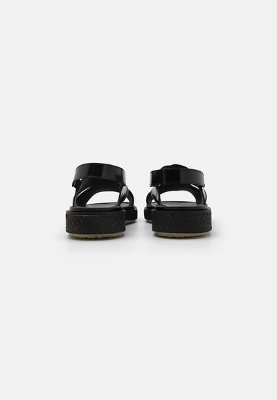 Sandales en cuir verni noir avec deux larges brides, dos ajustable et semelle en mousse noire texturée. Design plat, apparence minimaliste.
