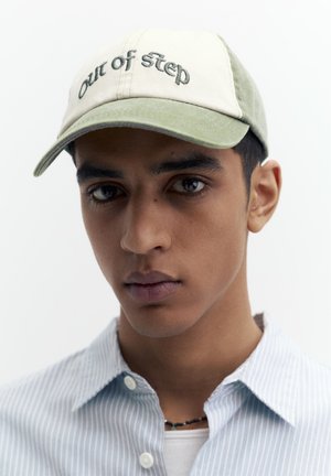 Mladić koji nosi bež i zelenu kapu s natpisom "Out of Step", prugastu svijetloplavu košulju i crnu ogrlicu od perli, neutralna pozadina.