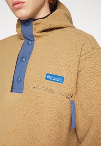 Pullover in pile color sabbia con patta blu a bottoni, patch del logo Columbia e tasca con zip. Tessuto testurizzato con un aspetto morbido e peloso.
