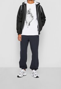 Zwarte lichtgewicht jas met een capuchon, witte graphic t-shirt met een handafdruk, marineblauwe joggingbroek en witte sportschoenen.