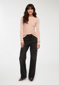 Jasnoróżowy sweter z teksturowanym splotem, zestawiony z czarnymi, szerokimi spodniami o wysokim stanie i brązowymi, spiczastymi szpilkami, stojący na tle w neutralnych kolorach.