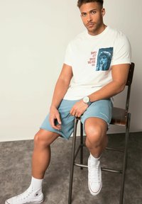 Bílá tričko s modrou grafikou a červeným nápisem, v kombinaci s modrými šortkami. Model má na sobě bílé tenisky a sedí na kovovém stoličce.