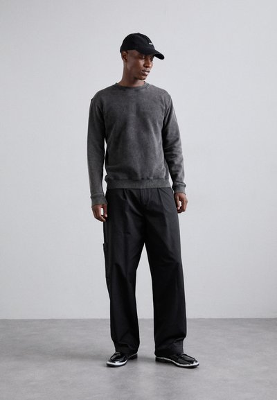 Felpa grigia scura con polsini a costine, pantaloni cargo neri con tasca laterale e cappellino nero. Le calzature presentano un design lucido e a contrasto.