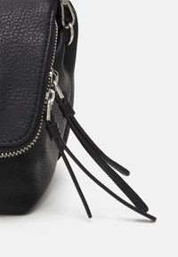 Coin de sac en cuir noir texturé avec deux fermetures éclair argentées visibles et de longues languettes en cuir noir sur fond blanc.