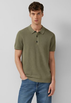 Młody mężczyzna o krótkich kręconych włosach, ubrany w oliwkowozieloną, krótkorękawową koszulkę polo i niebieskie dżinsy, stoi z jedną ręką w kieszeni.