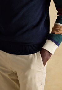 Person iført beige bukser med den ene hånd i lommen, en mørkeblå sweater og en flerfarvet stribet ærme synligt på armen.