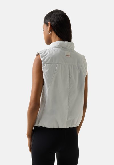 Kobieta o długich ciemnych włosach, nosząca srebrne kolczyki koła i białą pikowaną kamizelkę bez rękawów, zwrócona tyłem, w połączeniu z czarnymi legginsami.