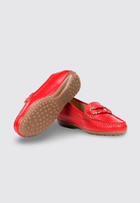 Par de mocasines de cuero rojo con suelas con tachuelas de goma y detalle de hebilla metálica en la parte frontal, exhibidos sobre un fondo gris claro.