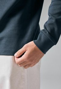 Mørkeblå langærmet skjorte med en glat tekstur. En hånd griber fat i kanten, hvilket afslører en lysere stof underneden. Simpelt design, ingen mønstre.