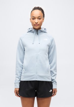 Kobieta o kręconych włosach nosi jasnoniebieską bluzę z kapturem na suwak i czarne szorty na tle jasnego, jednolitego tła.