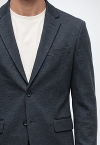 Homme portant un blazer gris foncé sur un t-shirt blanc à col rond sur un fond uni.
