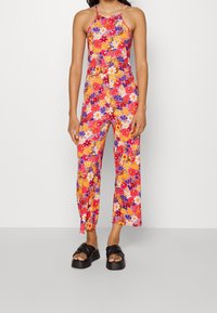 Mameluco floral con un patrón multicolor en rosa, naranja, púrpura y blanco. Presenta tirantes finos y un corte de pierna ancha, combinado con sandalias negras.