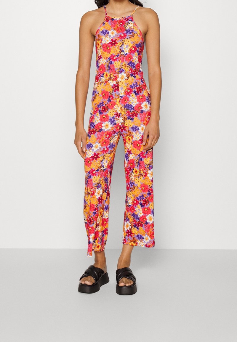 Mameluco floral con un patrón multicolor en rosa, naranja, púrpura y blanco. Presenta tirantes finos y un corte de pierna ancha, combinado con sandalias negras.