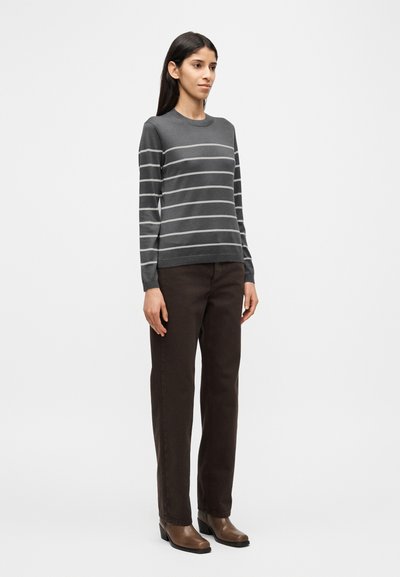 Γκρι πουλόβερ με μακριά μανίκια και οριζόντιες λευκές ρίγες, συνδυασμένο με σκουρό καφέ παντελόνι και καφέ μποτάκια αστραγάλου. Στέκεται σε απλό φόντο.