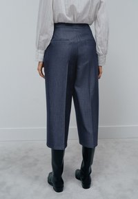 Personne portant un pantalon court taille haute, jambes larges, gris foncé, une chemise rayée claire et des bottes noires montantes, debout sur un tapis gris.