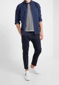 Man in een marineblauwe overhemd met knopen over een zwart-wit gestreept T-shirt, marineblauwe broek en witte sneakers met blauwe details.