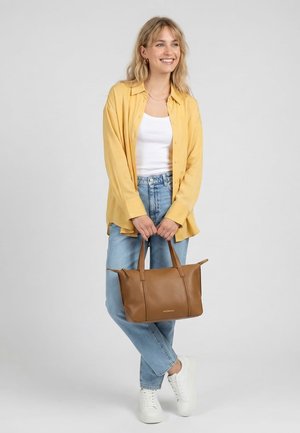 Glunderende vrouw in een geel shirt, wit topje, blauwe spijkerbroek en witte sneakers, die een bruine leren handtas vasthoudt tegen een effen lichte achtergrond.