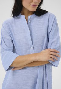 Camicia a righe blu e bianche con colletto a bande e chiusura con bottoni, caratterizzata da maniche corte e una vestibilità ampia. Texture di cotone morbido.