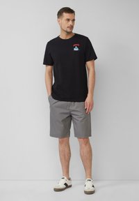 Schwarzes Baumwoll-T-Shirt mit rotem und blauem grafischem Design, kombiniert mit grauen Shorts und weißen Sneakers mit schwarzen Akzenten.