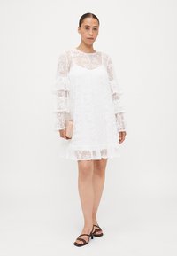 Valkoinen pitsimekko, jossa on pitkät, levenevät hihat, kerrokselliset röyhelöt helmassa, kukka-ommeltu ja rento malli. Lisänä pieni vaaleanpunainen clutch-laukku.