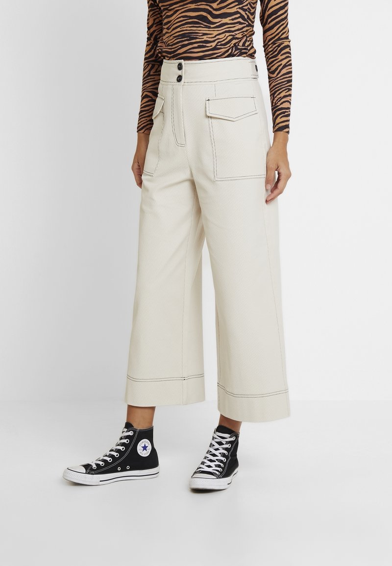 Personne portant un pantalon crème taille haute à jambes larges et coupé court avec de grandes poches avant, un haut à manches longues rayé comme un tigre, et des baskets montantes noires.