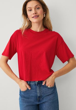 Kvinde med lysebrunt hår iført sølv hoop-øreringe, rød cropped top med korte ærmer og blå højtaljede jeans, hænderne i lommerne.