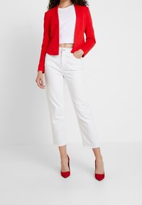 Rode cropped blazer over een witte, getailleerde top, gecombineerd met witte, hoge taille jeans. Het model draagt rode, puntige hakken. Gladde stof, gestructureerde pasvorm.