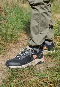 Chaussures de randonnée noires et grises avec accents beige, en mesh et matériaux synthétiques, dotées de lacets et d'une semelle robuste, posées sur un sentier en terre.