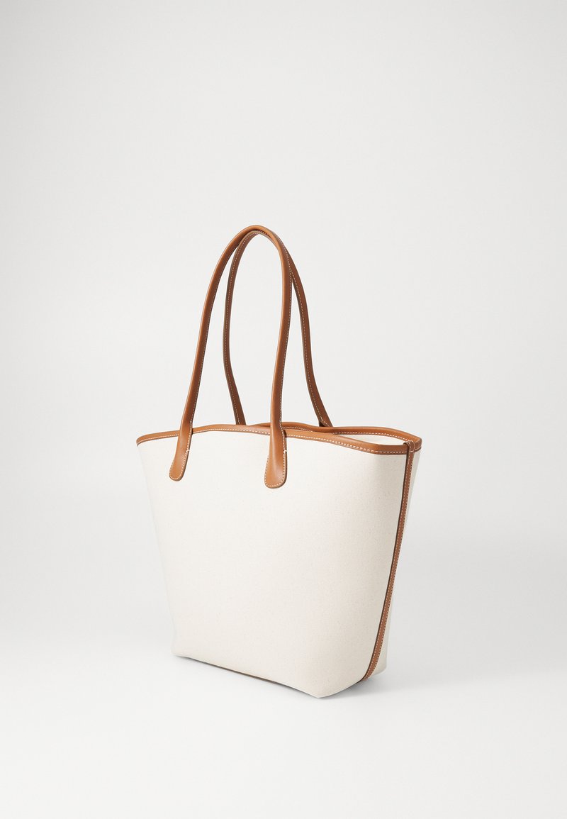 Sac en toile beige avec des poignées et des bordures en cuir tan. Présente un design minimaliste et une ouverture en haut, mettant en avant des lignes et des textures épurées.