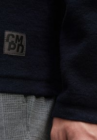 Navy strukturierter Stoff mit einem quadratischen schwarzen Etikett, auf dem "CMPD" am Saum gestickt ist. Angrenzende grau gemusterte Hosen sind teilweise sichtbar.