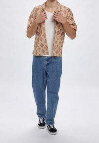 Koszula z krótkim rękawem w jasnobeżowym kolorze z czerwonymi geometrycznymi wzorami, noszona rozpięta na białym topie, zestawiona z niebieskimi dżinsami i czarnymi trampkami.