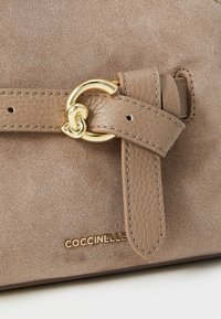 Beige suede handbag featuring a textured leather strap, gold-tone hardware, and embossed logo at the base. Closure has a distinct buckle detail.