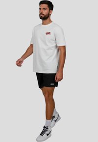 Camiseta blanca de algodón con un gráfico rojo, combinada con pantalones cortos negros. El modelo lleva zapatillas blancas y calcetines. Un look atlético simple y casual.