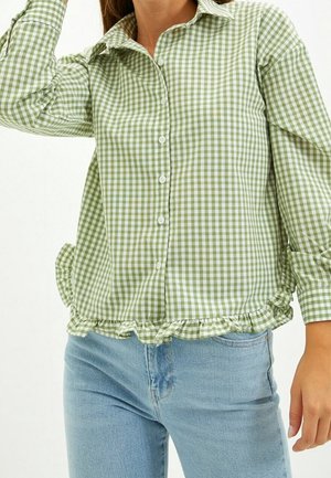 Woman wearing a green and white gingham button-up shirt with ruffled hem and light blue jeans, posing with arm raised.