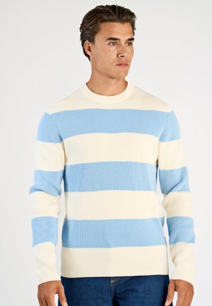 Uomo che indossa un maglione a coste con ampie strisce orizzontali color crema e azzurro chiaro, abbinato a jeans blu scuro, in piedi su sfondo bianco.