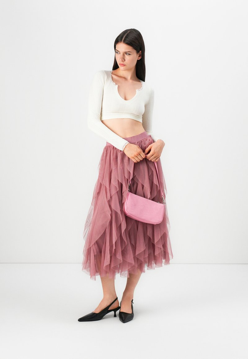 Weißes, langärmliges, kurzes Oberteil, geschichteter rosa Tüllrock, schwarze spitze Absatzschuhe und eine kleine rosa Handtasche mit glatter Textur.