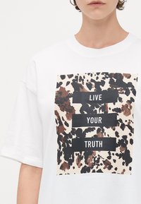 Person som bär en vit T-shirt med ett brun- och svartmönstrat kvadratiskt motiv och texten "LEVA DIN SANNING" i svarta block.