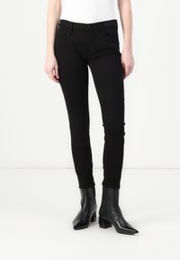 Jeans skinny negros con una textura suave, cintura de media altura, bolsillos delanteros y un cierre clásico con botón. Combinados con botas de tobillo negras y brillantes.