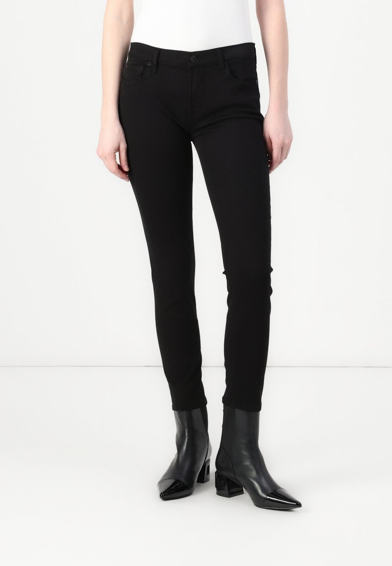 Jeans skinny negros con una textura suave, cintura de media altura, bolsillos delanteros y un cierre clásico con botón. Combinados con botas de tobillo negras y brillantes.