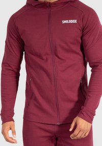 Sudadera con cremallera de color burdeos, hecha de una tela elástica, con un diseño ajustado, bolsillos laterales y un logo blanco en el pecho. Textura suave.