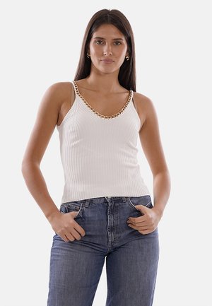 Donna con lunghi capelli scuri che indossa un top bianco senza maniche con dettagli in catena dorata e jeans di denim blu, in piedi con le mani nelle tasche.