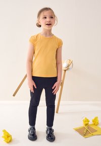 Gelbes Kurzarmoberteil mit gerafften Schultern, kombiniert mit schwarzen Leggings und glänzenden schwarzen Schuhen. Accessoires umfassen gelbes Papier und Schreibwaren.