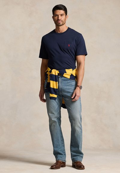 Mann iført marineblå t-skjorte, lyseblå jeans, brune sko, med gul og marineblå stripete genser bundet rundt livet, stående mot ensfarget bakgrunn.