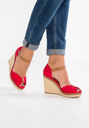 Rode sleehakken met een gedraaide stoffen design, open teen en een bruine enkelriem. Sleehak omwikkeld met jute voor extra textuur en contrast.